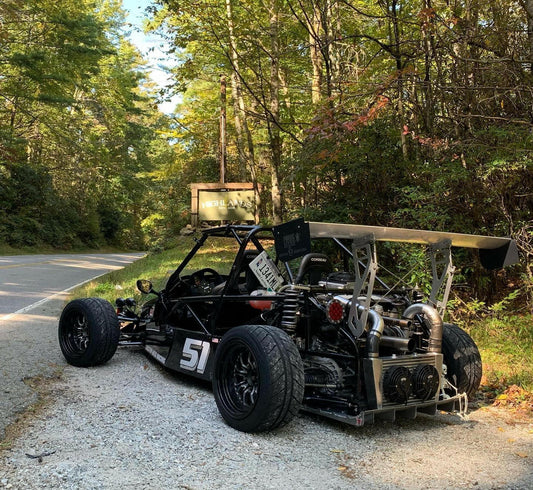 A custom-built, open-wheel race car, numbered 51, parked on a gravel pull-off beside a forested road. The car features a DF Goblin Big Wang Kit by 9livesracing for maximum downforce and intricate engine components visible. Trees and foliage frame the scene, with a road sign partially visible in the background.