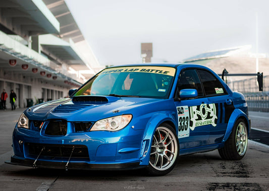 A blue Subaru Impreza STI Big Wang kit '00-07 GD from 9livesracing, featuring a black front splitter and a large aluminum rear wing, is parked on the track. It boasts numerous racing decals, including "Super Lap Battle" on the windshield and "333" on the side. The background reveals a bustling racing circuit and grandstands, emphasizing its high-octane downforce design.