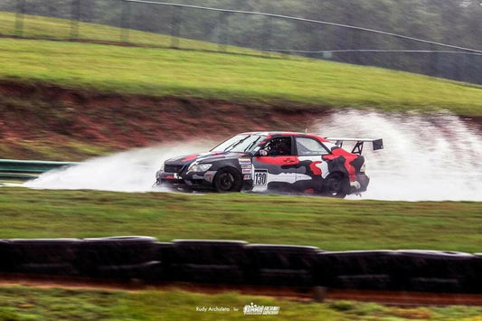 A race car, labeled with the number 100, zips through a wet track at high speed, creating a large spray of water behind it. Sporting a black and red camouflage livery and making precise down-force adjustments, the Is300 Big Wang Kit 98-04 xe10 by 9livesracing navigates a turn with grassy areas and tire barriers in the background.