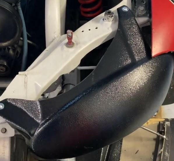A close-up view of the Miata Headlight Blanks NB1-Only, a black, textured metal component from Nine Lives Racing, reveals its attachment to a white frame with screws. A red coiled spring is visible in the background, along with other metallic elements and partially visible robust plastic components that contribute to its durability.