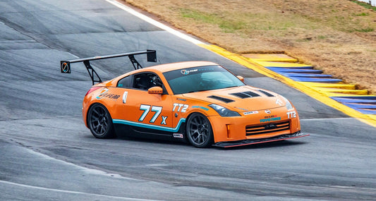 An orange sports car with the number 77X and T2 decals speeds around a racetrack, flaunting its 9livesracing 350z Big Wang Kit '02-08 Z33 performance. The car features extruded aluminum wings, black wheels, and various sponsor stickers. The track is gray with a yellow and blue striped barrier on the side, and grass is visible in the background.