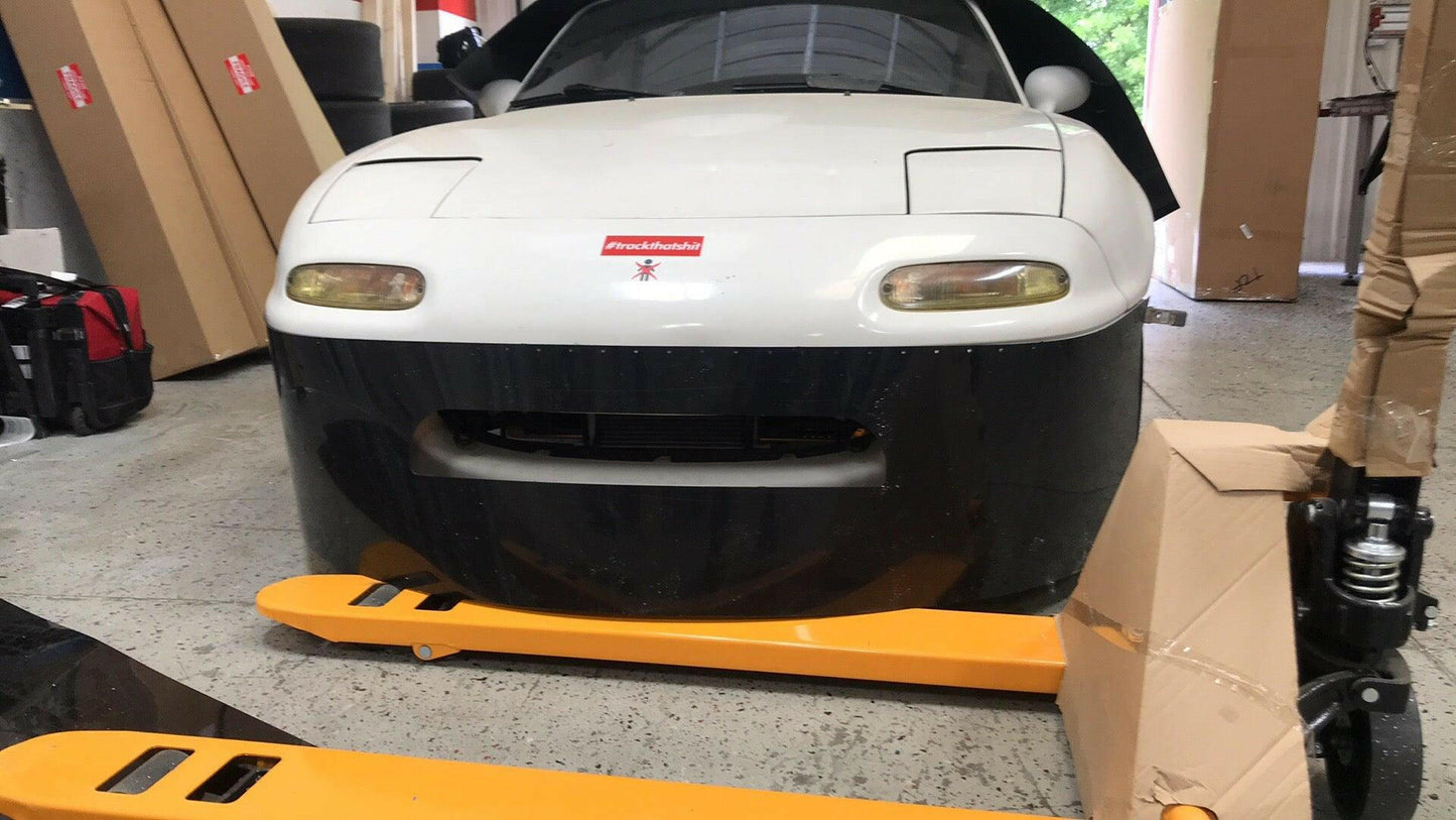 A white and black Mazda Miata with a detailed front end is parked indoors. A yellow pallet jack is positioned in front of the car, and various boxes and equipment are scattered nearby. The car's hood is open, revealing part of the engine, while a 9livesracing Miata Damn Dam '90-05 NA/NB suggests enhanced downforce capabilities.