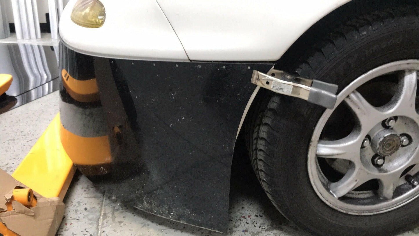 A close-up of the front left wheel and bumper of a Mazda Miata featuring the 9livesracing Miata Damn Dam '90-05 NA/NB. The black bumper is partially detached, revealing silver hardware and a gap that exposes the air dam. The silver-gray wheel displays visible tread and detailing, while an orange and black floor jack is partially visible on the left.