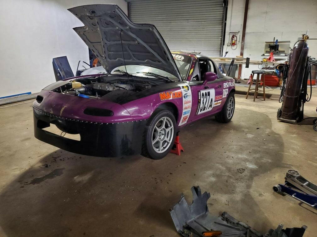 A purple race car with number 337 under the hood sits in the garage, supported on jack stands. Various tools and parts are scattered on the floor around it, including a 9livesracing Miata Damn Dam '90-05 NA/NB air dam, while industrial equipment and a partially closed rolling door loom in the background.