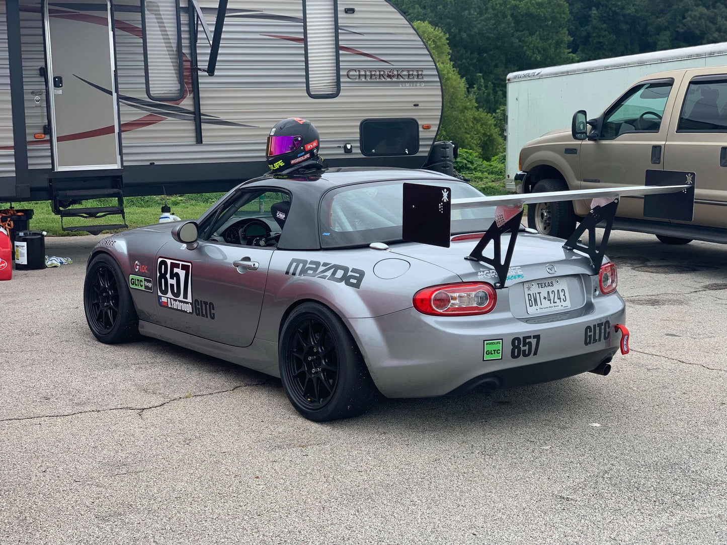 A gray race car, labeled with number 857, is parked in front of an RV and a van in a paddock area. The car features a Miata Big Wang Kit '06-15 NC *V1* by 9livesracing, black wheels, and racing decals, including a Mazda logo. A helmet rests on the car's roof.