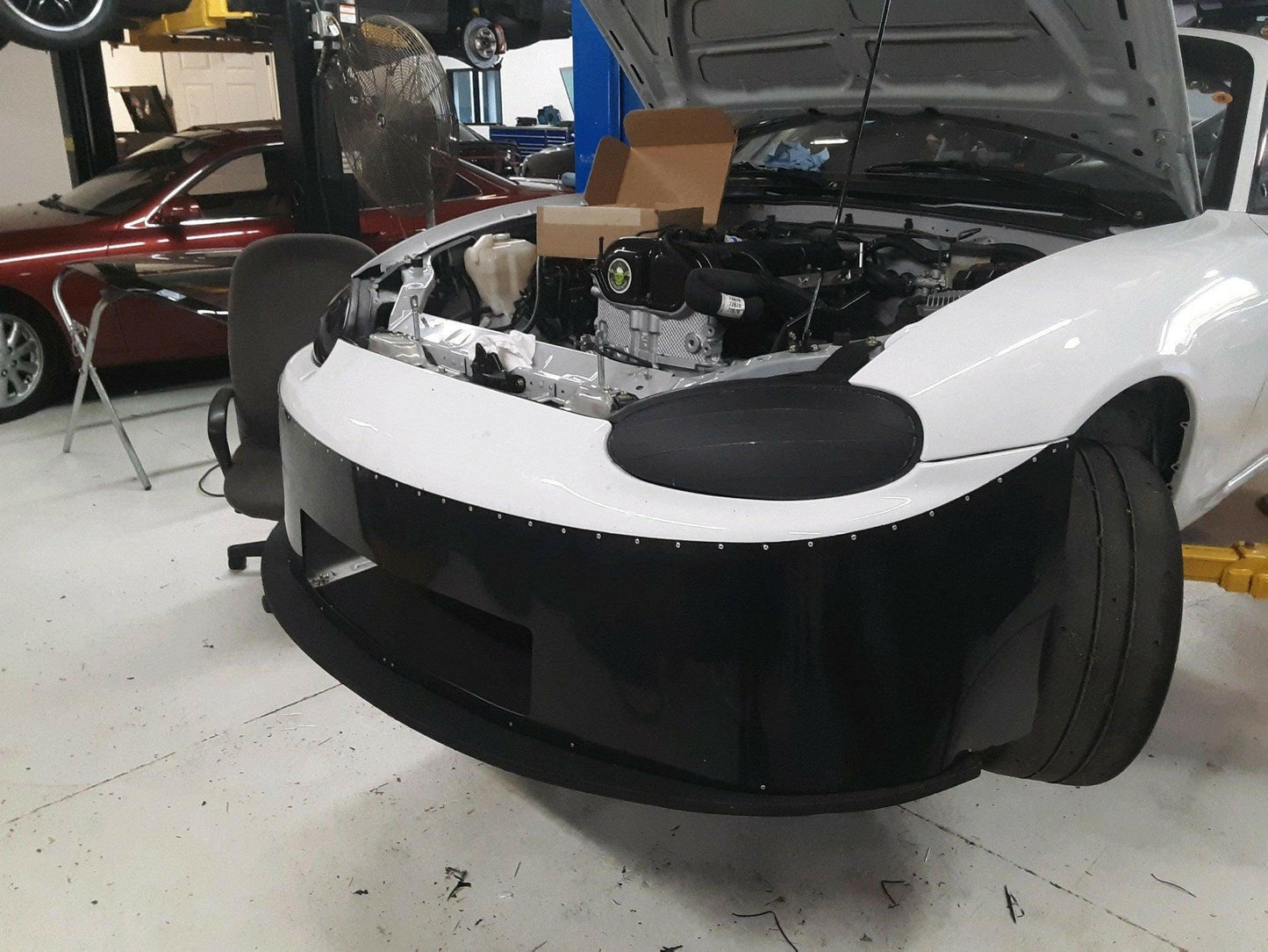 A white Mazda Miata with its hood open is undergoing maintenance in a garage. The front bumper and the Miata Damn Dam '90-05 NA/NB from 9livesracing have been removed, exposing the engine bay. A chair with tools and parts sits to the left, while another car is elevated on a lift in the background.