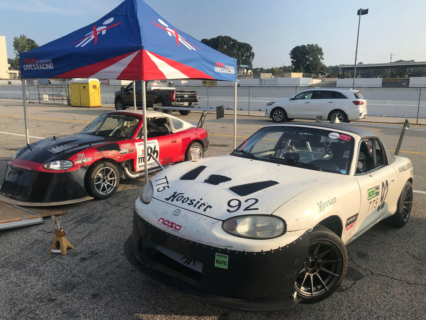 Two race cars are parked under a canopy adorned with a British flag design and the text "Weiss Racing." One car is red and black, while the other is white with the number "92" on its hood. Both cars are equipped with Miata Damn Dam '90-05 NA/NB by 9livesracing, featuring large front splitters and air dams to maximize downforce. A white SUV is parked nearby, and a race track fence can be seen in the background.