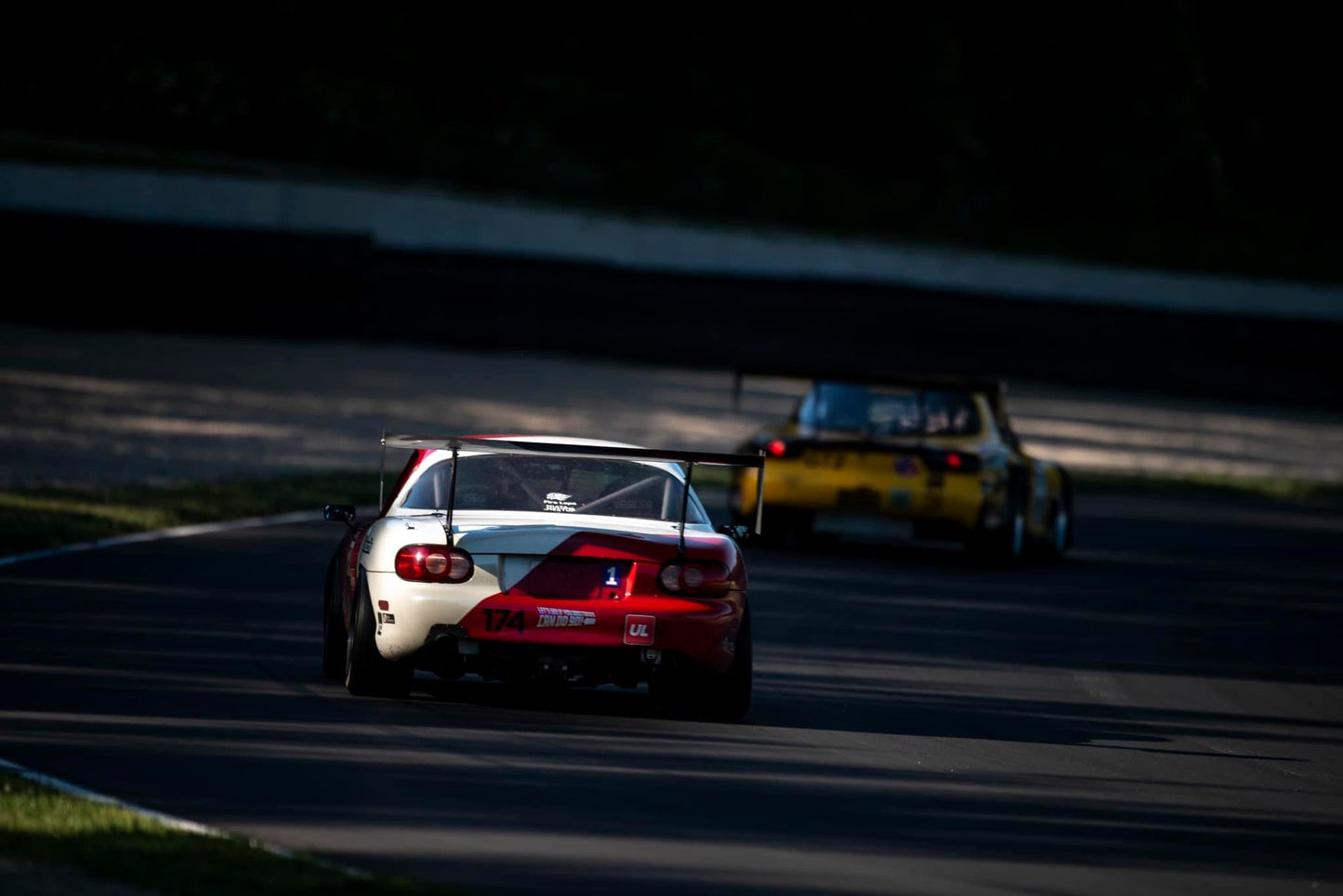 Two race cars speed down a darkened track, with sunlight highlighting parts of the road. The car in the foreground, outfitted with a 9livesracing Miata Big Wang Kit '90-05 NA/NB for impressive down-force, is red and white with the number 174. In the background, an American-made yellow car adds to the thrill on this professional racing circuit.