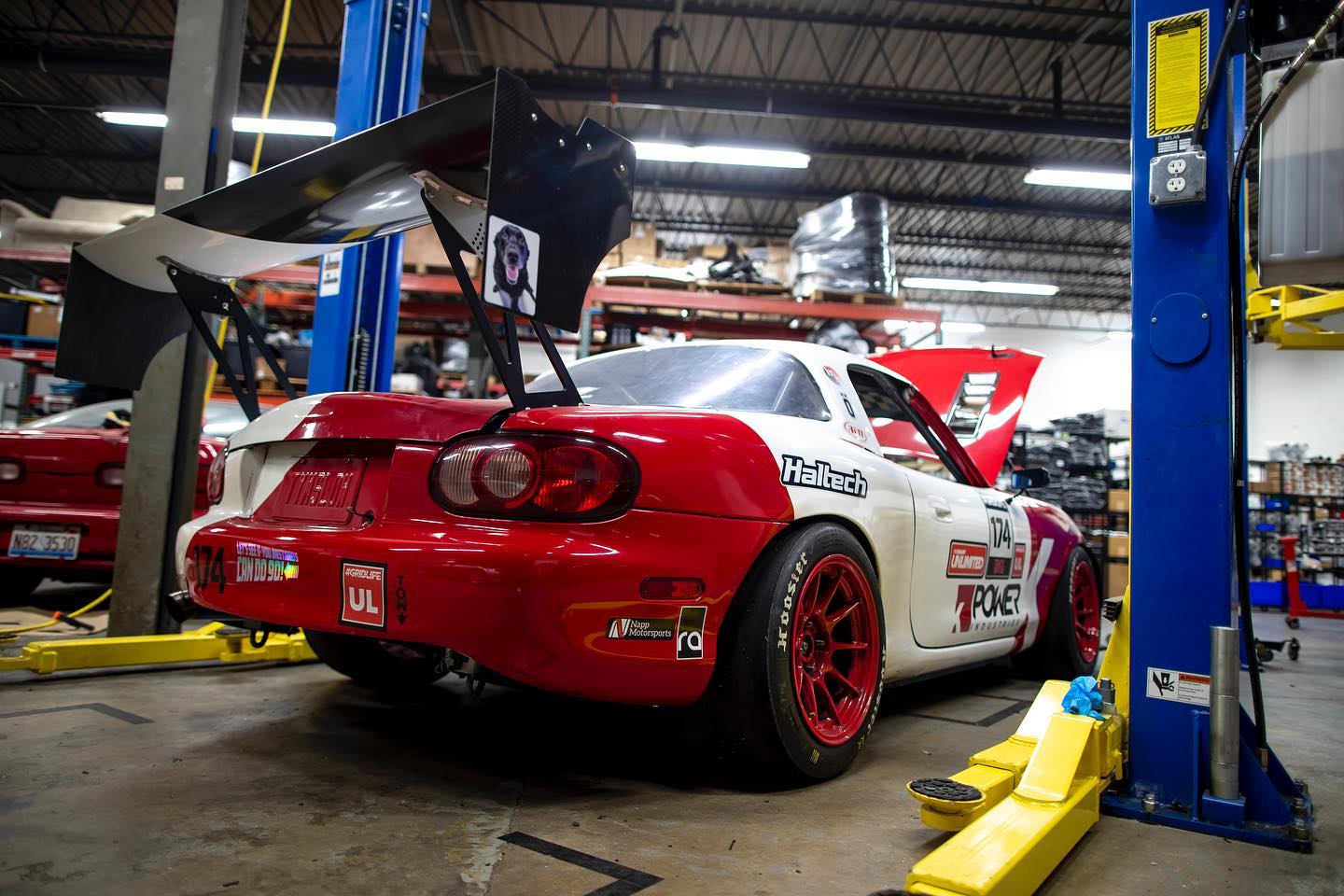 A red and white racing car equipped with the 9livesracing Miata Big Wang Kit '90-'05 NA/NB is elevated on a hydraulic lift in an American-made workshop garage. The car boasts a large rear wing for increased downforce and red rims. Various tools and parts are visible in the background, with shelves and other equipment scattered throughout the space.