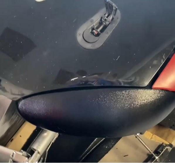 Close-up view of the Miata Headlight Blanks NB1-Only by Nine Lives Racing on a black vehicle, showing part of the mounting area crafted from robust plastic and an attached wiring component. The surrounding area includes reflections and portions of the car's body, highlighting the vehicle's glossy finish.