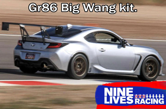 A silver sports car on a racetrack, equipped with a large rear wing made from extruded aluminum wings, is captured in motion. "GR86 / BRZ Wang Kit '22+" is text at the top. The bottom right corner features a "9livesracing" logo with a red and blue design.