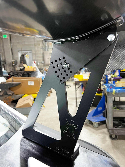Close-up of a black metal bracket mounted on a surface, with a workshop in the background. The bracket has a perforated area and features the 9livesracing logo with a small ant. Amidst scattered tools and equipment, you notice parts of what seems to be an ambitious BMW M2 CS Racing Big Wang Kit project underway.