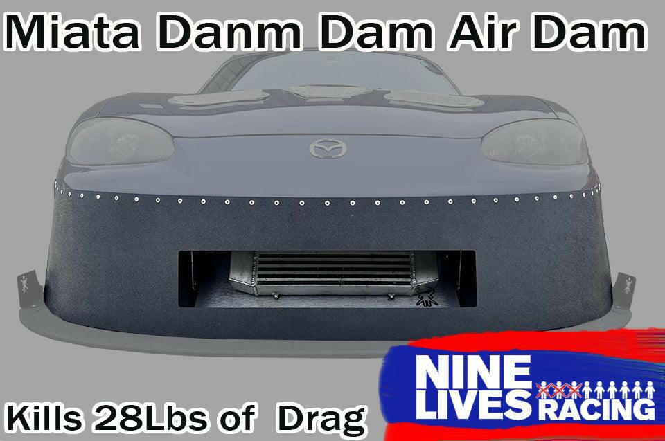 A front view of a grey Mazda Miata with an aftermarket air dam attached. Text at the top reads "Miata Damn Dam '90-05 NA/NB" and at the bottom reads "Kills 28Lbs of Drag". The 9livesracing logo is in the lower right corner, showcasing enhanced downforce.