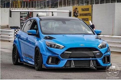 A bright blue Ford Focus RS is parked near a race track, showcasing a modified front bumper and aerodynamic enhancements. The car features black wheels, a lowered stance, and a prominent 9livesracing Focus/RS Big Wang Kit '11-18 3rd Gen aluminum wing for added down-force. Industrial and environmental service signs are visible in the background.