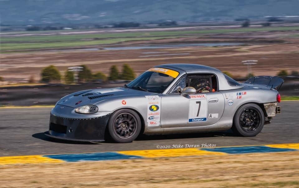 A silver race car with the number 7 on its doors speeds along a racetrack during a race, equipped with a Miata Damn Dam '90-05 NA/NB by 9livesracing for added downforce. The car, possibly inspired by Mazda Miatas, is adorned with various sponsor stickers. The background features fields and hills under a blue sky. Photo by Mike Doran, 2020.