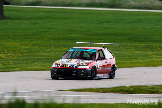 A red Civic Hatch EF Wang kit 88-91, equipped with various colorful stripes and a large rear wing reminiscent of the Big Wang GT3 from 9livesracing, races on a track surrounded by green grass. The car's hood features multiple sticker designs. The track curves slightly, and the background is blurred, indicating motion and impressive down-force.