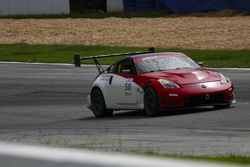 A red and white race car with the number 50 on the door speeds around a racetrack, equipped with a 9livesracing 350z Big Wang Kit '02-08 Z33, featuring GT3 performance and extruded aluminum wings. The car boasts a prominent rear spoiler and various sponsor logos. The track is lined with grass and barriers, with tire walls and gravel in the background.