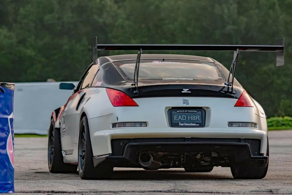 Rear view of a white sports car equipped with the 350z Big Wang Kit '02-08 Z33 from 9livesracing, showcasing extruded aluminum wings and black detailing. The car sports a license plate reading "EAD H8R" and is parked on a concrete surface with greenery in the background.