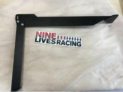 A black metal bracket with bolts is placed on a light-colored surface. Beneath the bracket, there is a sticker that reads "9LIVESRACING" in bold letters, with small human figures representing the "I" in "LIVES." These brackets are ideal for downforce adjustments and performance airfoils on Impreza Wagon Big Wang kit '00-07 GG from 9livesracing.