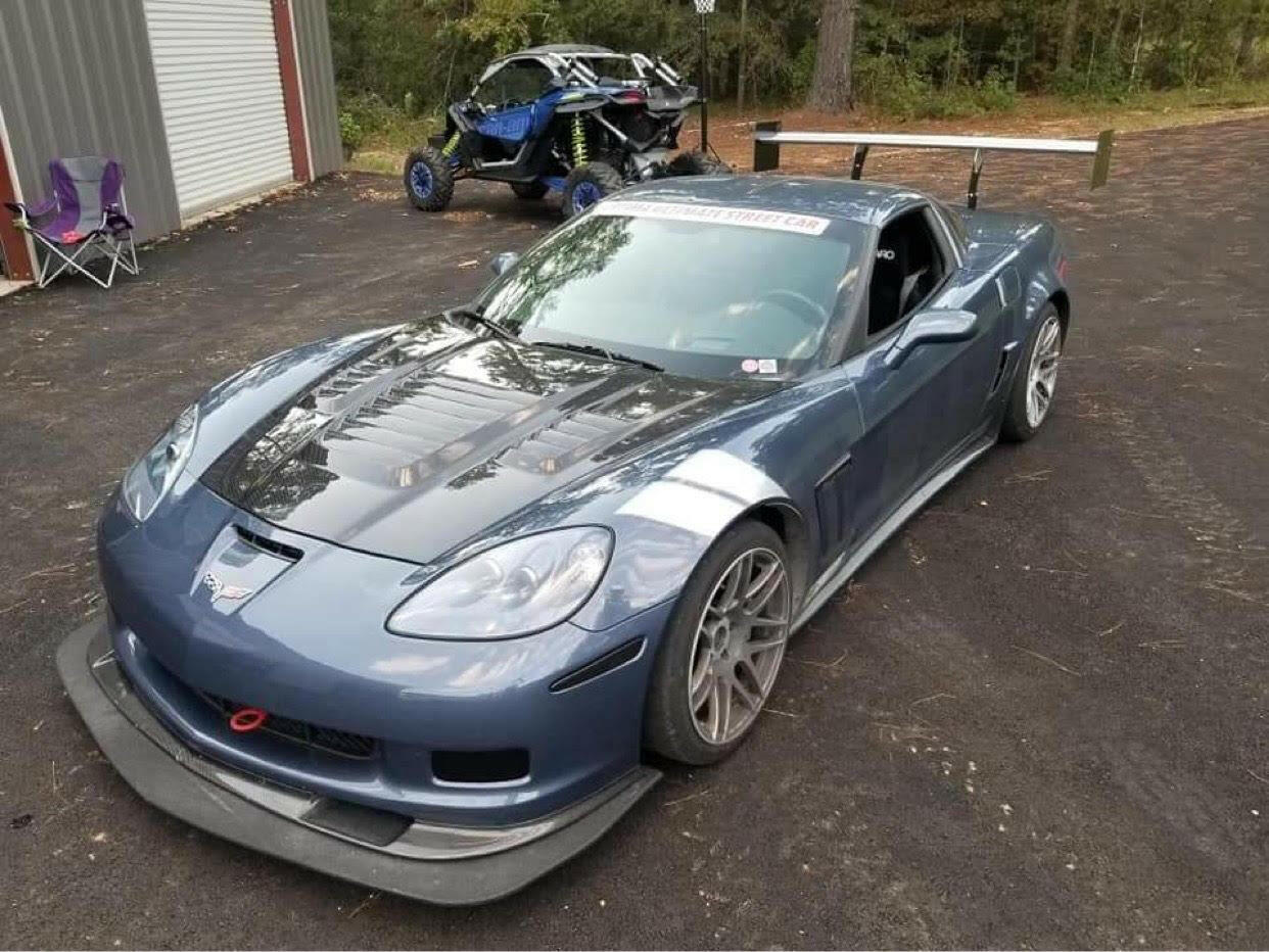 A Corvette fitted with a 9livesracing C6 & C5 '05-'13 splitter, featuring a carbon fiber hood, low friction polymer aerodynamic modifications, and sporting vibrant racing decals is parked on an asphalt surface. In the background, there is a blue off-road vehicle near a building with a partially open garage door, and a purple folding chair is positioned nearby.