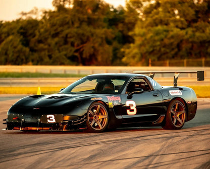 A sleek black race car marked with the number 3 is parked on a race track. The car features various sponsor decals, red-accented wheels, and a Corvette Big Wang Chassis Mount ’97-13 C5/6 rear spoiler by 9livesracing designed to optimize lift-to-drag ratio. Behind it, there is green foliage and a hint of a sunset sky.