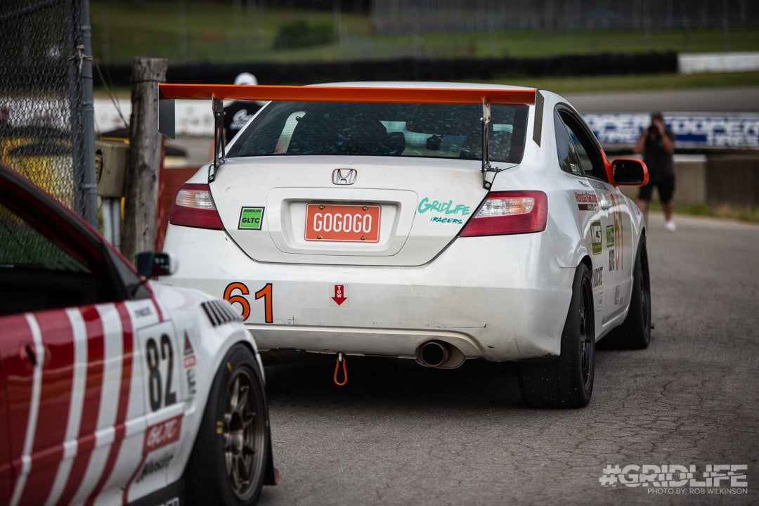 A white car displaying the number 61 and equipped with an orange spoiler, featuring the Civic Coupe Wang kit '06-11 from 9livesracing for an optimal lift-to-drag ratio, has a license plate reading "GOGOGO." Positioned on a racetrack next to another car numbered 82, a person stands near the track. The image is branded with "#GRIDLIFE.