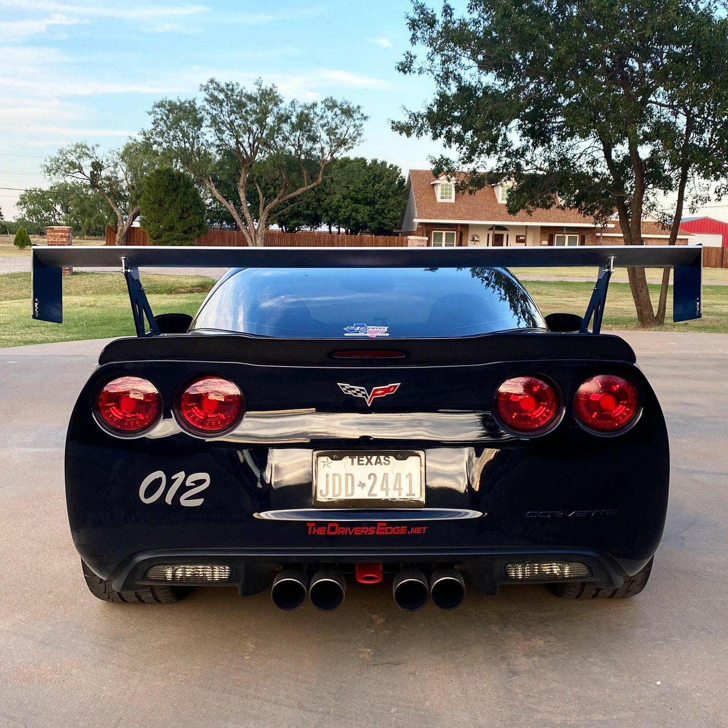 A black Corvette equipped with the 9livesracing Corvette Big Wang Trunk Mount Kit '97-13 C5/6 and red taillights is parked on a driveway. The license plate reads "JDD-2441" and is from Texas. The car has the number "012" on the back, with trees and a house visible in the background.