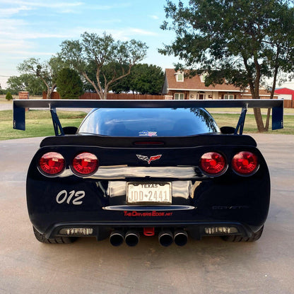 A black Corvette equipped with the 9livesracing Corvette Big Wang Trunk Mount Kit '97-13 C5/6 and red taillights is parked on a driveway. The license plate reads "JDD-2441" and is from Texas. The car has the number "012" on the back, with trees and a house visible in the background.