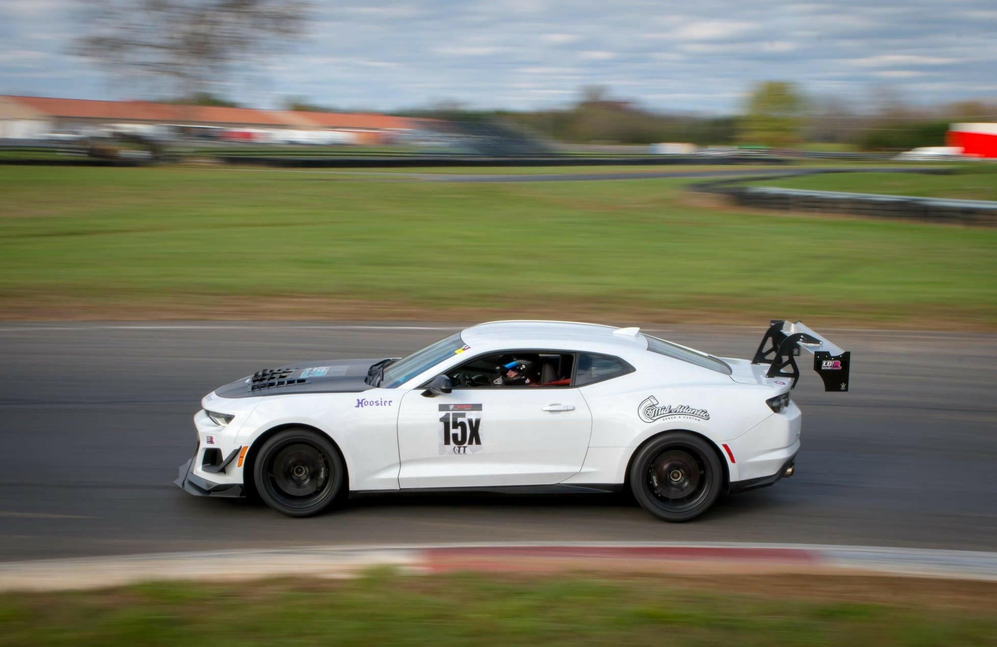 A white "15X" Camaro Wang 2016+ 6th Gen from 9livesracing speeds on the track during a daytime race, equipped with aluminum wings for optimized downforce adjustments and a prominent rear wing. Blurred grassy areas and a building in the background hint at its GT3 performance and swift motion.