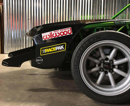 Close-up of the rear left side of a race car featuring the Wilwood Disc Brakes and Racepak stickers. The car has a black body, exposed green framework, and a tire marked with "VICRA V7" on a silver alloy rim. An impressive Exocet Chrome Horn Nose Wang Kit by 9livesracing adds to its aerodynamic prowess against the backdrop of a corrugated metal wall.