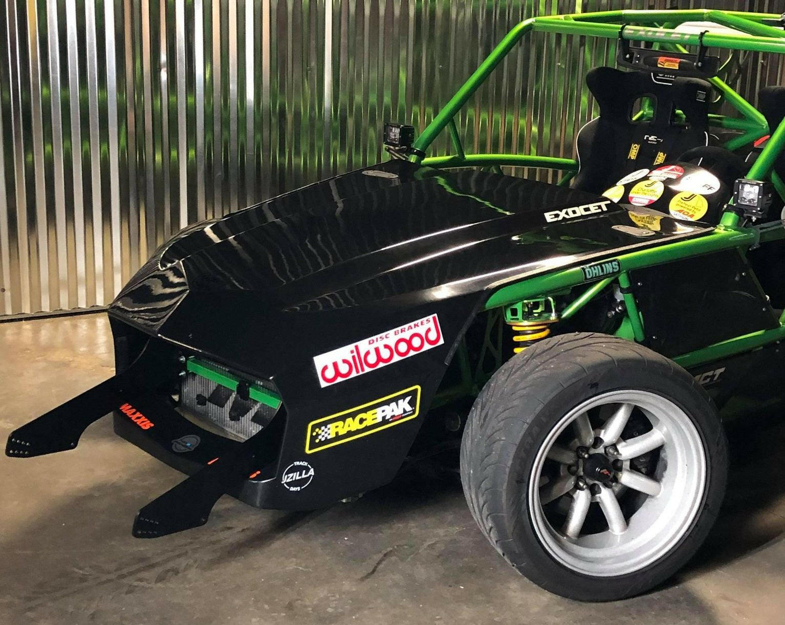 An Exocet Chrome Horn Nose Wang Kit by 9livesracing: a custom-built, exposed-frame race car with a glossy black hood, green roll cage, and various sponsor decals. It features aluminum wings for enhanced aerodynamics and has a low-profile design with no headlights. The detailed coil suspension is visible as the car is parked by a corrugated metal wall.
