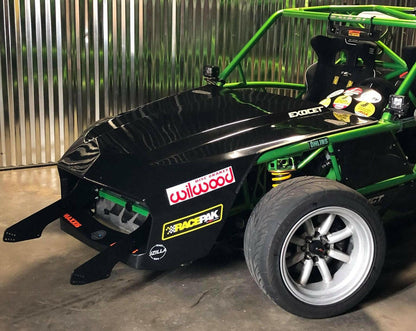 An Exocet Chrome Horn Nose Wang Kit by 9livesracing: a custom-built, exposed-frame race car with a glossy black hood, green roll cage, and various sponsor decals. It features aluminum wings for enhanced aerodynamics and has a low-profile design with no headlights. The detailed coil suspension is visible as the car is parked by a corrugated metal wall.
