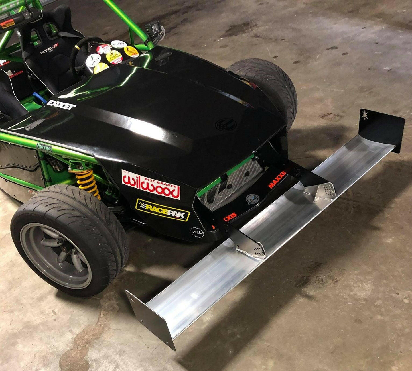 A close-up of a custom-built race car showcases its sleek, partially black and green exterior. The car features an Exocet Chrome Horn Nose Wang Kit by 9livesracing, visible decals from various brands, high-performance tires, yellow suspension coils, and aluminum wings. It is parked indoors on a concrete floor.