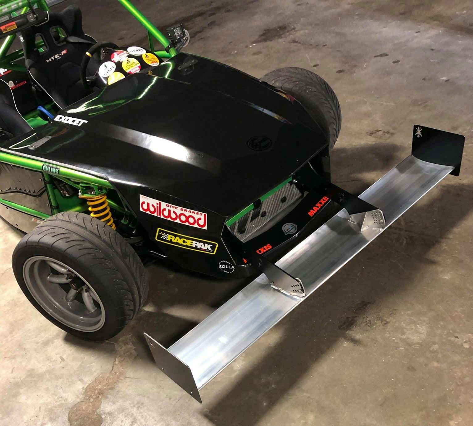 A close-up of a custom-built race car showcases its sleek, partially black and green exterior. The car features an Exocet Chrome Horn Nose Wang Kit by 9livesracing, visible decals from various brands, high-performance tires, yellow suspension coils, and aluminum wings. It is parked indoors on a concrete floor.