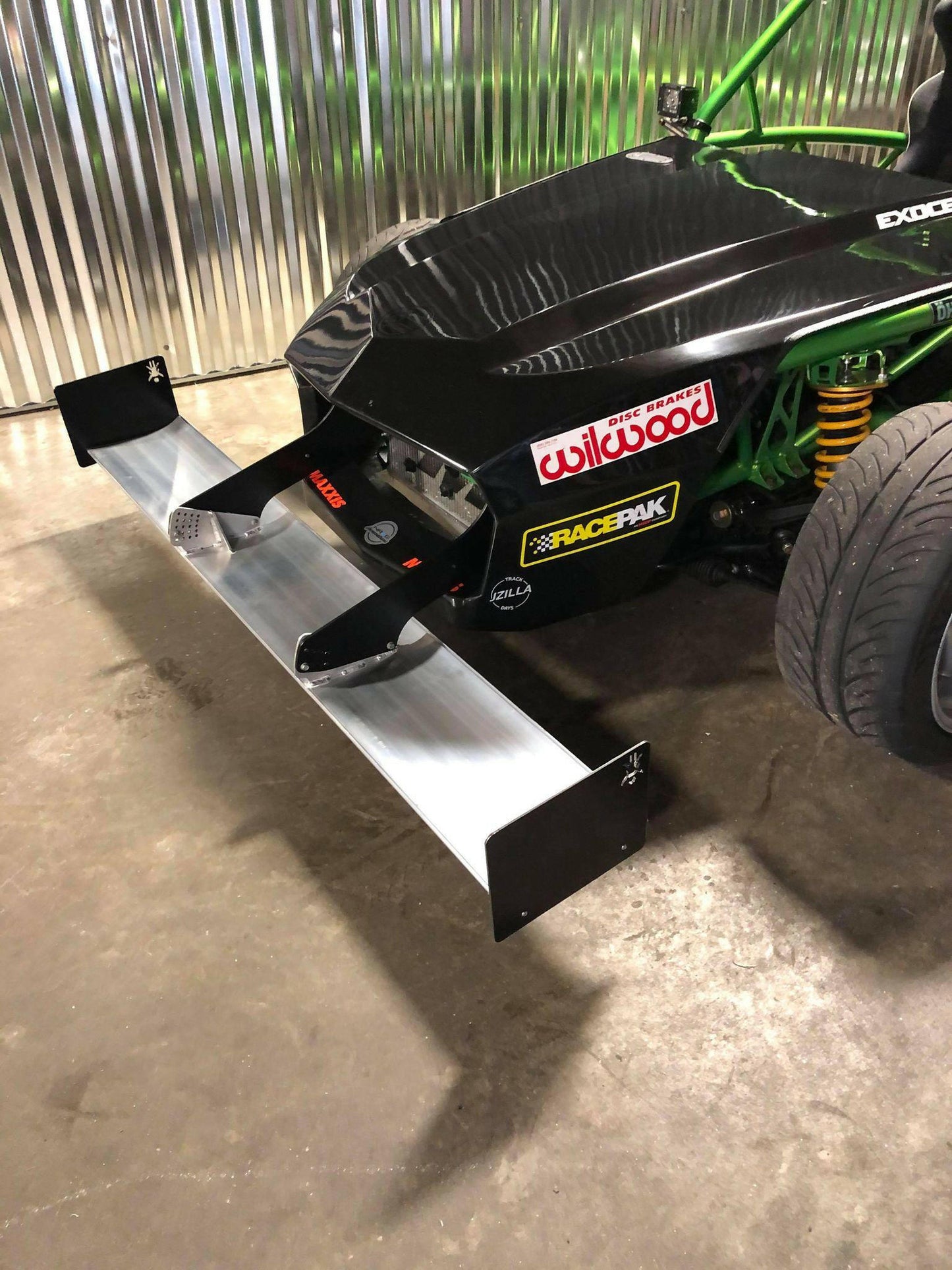 A close-up view of a black race car featuring various sponsor stickers on its front, including "Wilwood" and "RacePak." The vehicle is equipped with the 9livesracing Exocet Chrome Horn Nose Wang Kit, whose metal front splitter perfectly complements the aluminum wings and green roll cage, along with the visible suspension components.