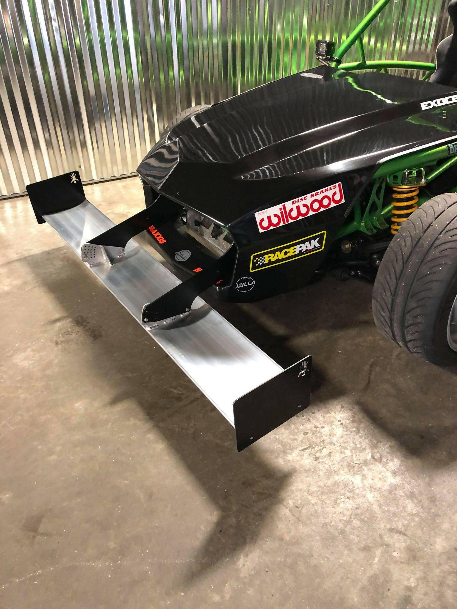 A close-up view of a black race car featuring various sponsor stickers on its front, including "Wilwood" and "RacePak." The vehicle is equipped with the 9livesracing Exocet Chrome Horn Nose Wang Kit, whose metal front splitter perfectly complements the aluminum wings and green roll cage, along with the visible suspension components.