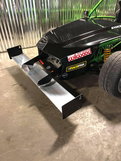 A close-up view of a black race car featuring various sponsor stickers on its front, including "Wilwood" and "RacePak." The vehicle is equipped with the 9livesracing Exocet Chrome Horn Nose Wang Kit, whose metal front splitter perfectly complements the aluminum wings and green roll cage, along with the visible suspension components.