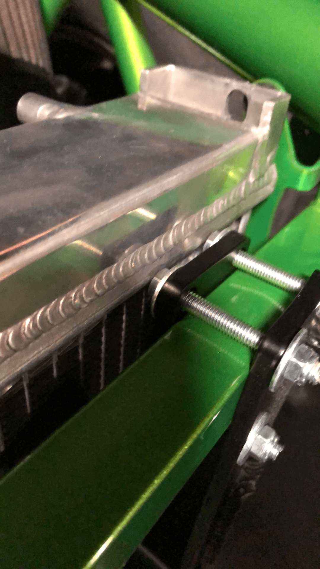 Close-up image of the Exocet Chrome Horn Nose Wang Kit by 9livesracing, showing bolts attaching it to a green structure, possibly part of a vehicle or machinery. The focus is on the details of the bolts and metal parts, highlighting the construction and assembly, reminiscent of precision found in aluminum wings.