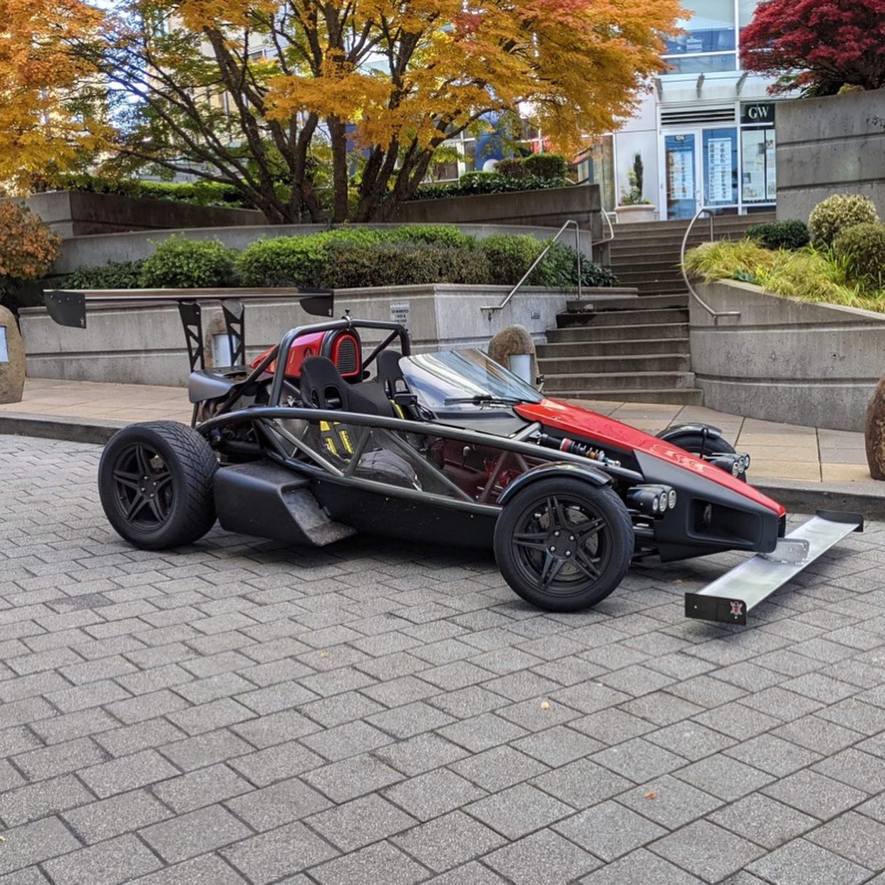 The Ariel Atom Big Wang by 9livesracing, a striking black and red open-wheel sports car, is parked on a paved area surrounded by trees adorned with autumn foliage. This vehicle features a unique, lightweight design with exposed wheels, a low seating position, and sleek airfoils that enhance its lift-to-drag ratio. In the background, there is a staircase and building visible.