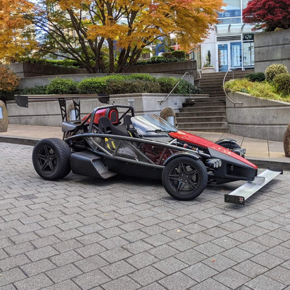 The Ariel Atom Big Wang by 9livesracing, a striking black and red open-wheel sports car, is parked on a paved area surrounded by trees adorned with autumn foliage. This vehicle features a unique, lightweight design with exposed wheels, a low seating position, and sleek airfoils that enhance its lift-to-drag ratio. In the background, there is a staircase and building visible.