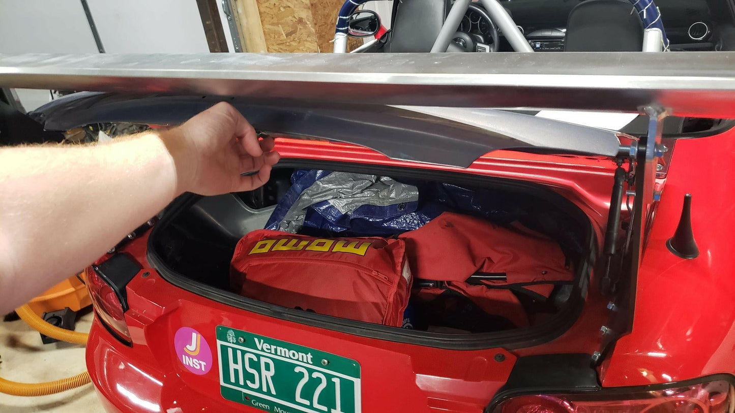 A person's hand lifts the Miata Big Wang Kit '06-15 NC (V2) rear spoiler from 9livesracing, enhancing the racing aerodynamics of a red sports car. The open trunk reveals various items, including a red MOMO-branded bag. Sporting a Vermont license plate labeled "HSR 2211," the car is parked in a garage.