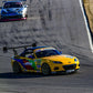 A yellow Mazda MX5 Splitters 06-15 NC by 9livesracing, sporting the number 55 and adorned with vibrant graphics and sponsors' logos, leads on a curved racetrack. It features a large rear wing and the sleek Rocket Nose V2 design, while being closely followed by a silver car. The background reveals a paved track bordered by grassy areas.