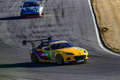 A yellow Mazda MX5 Splitters 06-15 NC by 9livesracing, sporting the number 55 and adorned with vibrant graphics and sponsors' logos, leads on a curved racetrack. It features a large rear wing and the sleek Rocket Nose V2 design, while being closely followed by a silver car. The background reveals a paved track bordered by grassy areas.