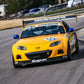 A yellow Mazda MX5 Splitters 06-15 NC, branded by 9livesracing and equipped with a Rocket Nose V2, speeds along a curved track. The car sports various sponsor decals, including Snap-on and Hankook. The driver is not visible in the image, and the background features a grassy area as well as several buildings.
