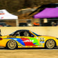 A colorful 9livesracing Mazda MX5 Splitters 06-15 NC, featuring the number 55, is speeding on a racetrack. Adorned with vibrant rainbow-themed paint and various sponsor logos, its Rocket Nose V2 cuts through the air. In the background, tents, a few people, and some racing equipment complete the lively scene.