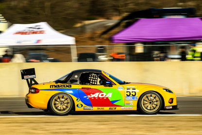 A colorful 9livesracing Mazda MX5 Splitters 06-15 NC, featuring the number 55, is speeding on a racetrack. Adorned with vibrant rainbow-themed paint and various sponsor logos, its Rocket Nose V2 cuts through the air. In the background, tents, a few people, and some racing equipment complete the lively scene.