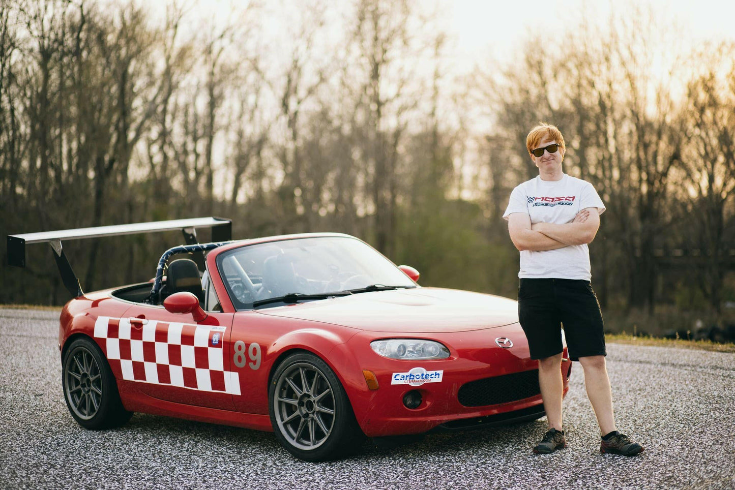 A person wearing a white t-shirt, black shorts, and sunglasses stands with arms crossed beside a red convertible sports car featuring several racing stickers, a 9livesracing Miata Big Wang Kit '06-15 NC (V2) for optimal down-force, and the number 89. The car displays a checkerboard design on the side. Trees are visible in the background.