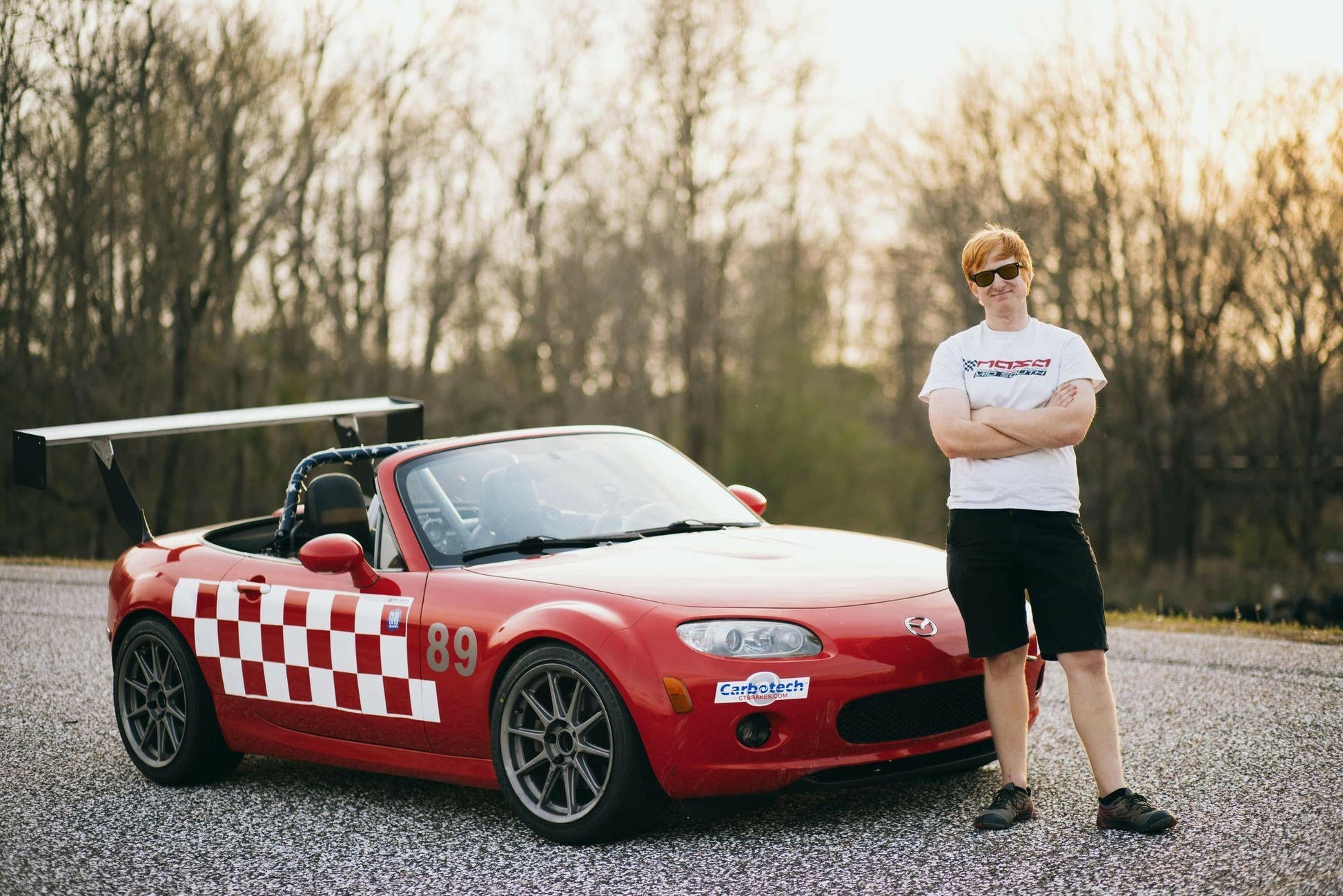 A person wearing a white t-shirt, black shorts, and sunglasses stands with arms crossed beside a red convertible sports car featuring several racing stickers, a 9livesracing Miata Big Wang Kit '06-15 NC (V2) for optimal down-force, and the number 89. The car displays a checkerboard design on the side. Trees are visible in the background.