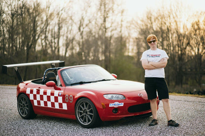 A person wearing a white t-shirt, black shorts, and sunglasses stands with arms crossed beside a red convertible sports car featuring several racing stickers, a 9livesracing Miata Big Wang Kit '06-15 NC (V2) for optimal down-force, and the number 89. The car displays a checkerboard design on the side. Trees are visible in the background.