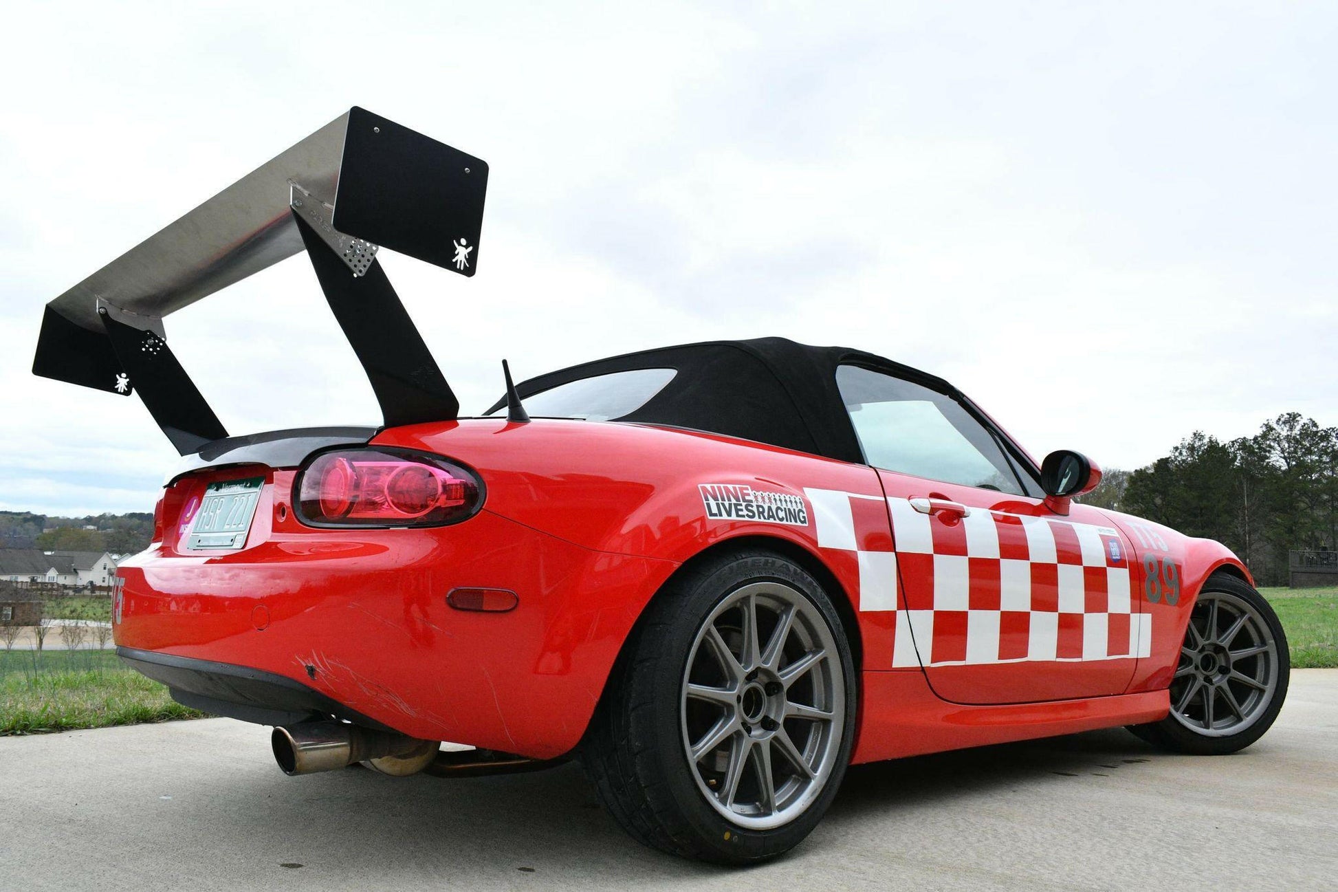 A red sports car featuring a Miata Big Wang Kit '06-15 NC (V2) from 9livesracing is shown parked on a concrete surface. The car boasts a black convertible top, white checkerboard pattern on the side, and labels from a racing team, enhancing its racing aerodynamics with the large performance wing spoiler. Trees and a cloudy sky can be seen in the background.
